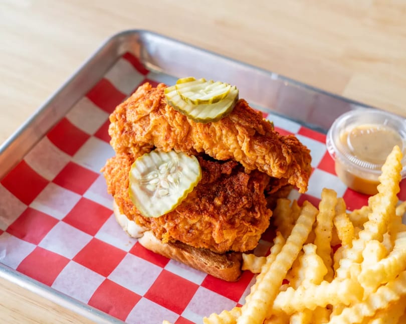 Crispy fried chicken sandwich with pickles, crinkle-cut fries, and dipping sauce on a red checkered tray.