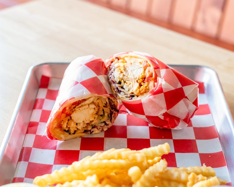 Two chicken wraps in red and white checkered paper, with crinkle-cut fries on a metal tray.