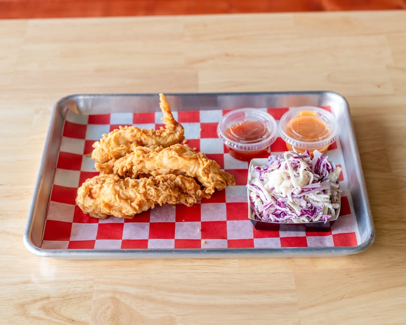 Crispy fried chicken tenders, coleslaw, and two dipping sauces on a metal tray with checkered paper.