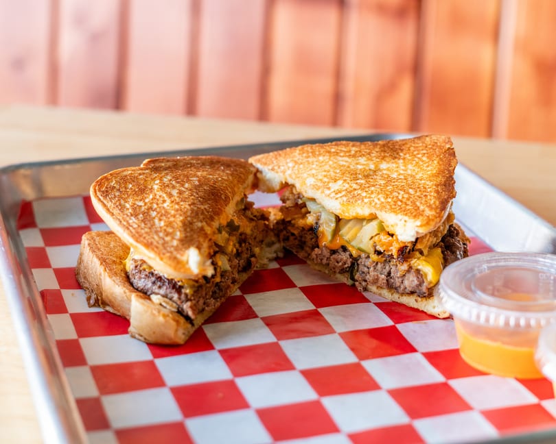 A grilled cheese burger, cut in half, on a red and white checkered paper-lined tray.