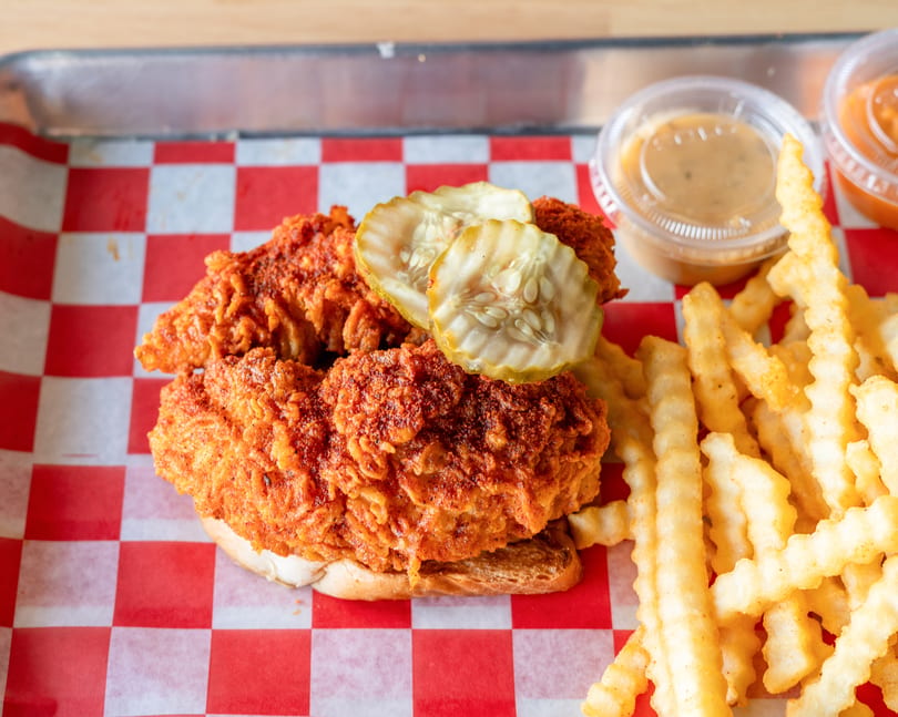 Spicy fried chicken sandwich with pickles and crinkle-cut fries on red and white checkered paper.