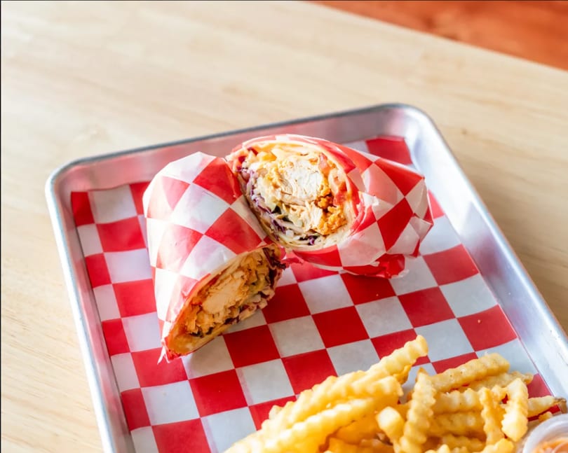 A chicken wrap cut in half, wrapped in red and white checkered paper, with crinkle-cut fries.