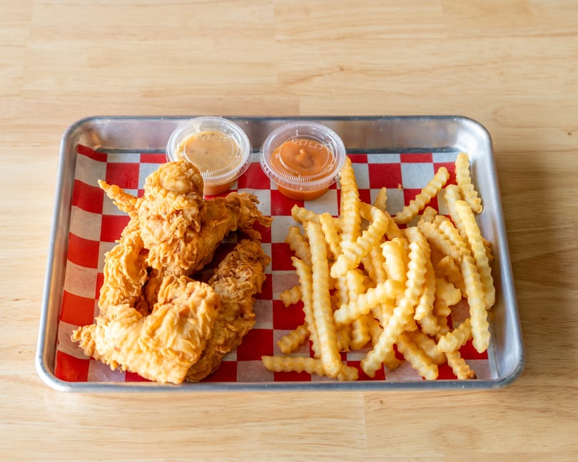 Fried chicken tenders and crinkle-cut fries with two dipping sauces on a metal tray.
